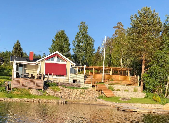 Hus vid sjökanten med röda markiser, stor träterrass, brygga och grönskande träd runtomkring.