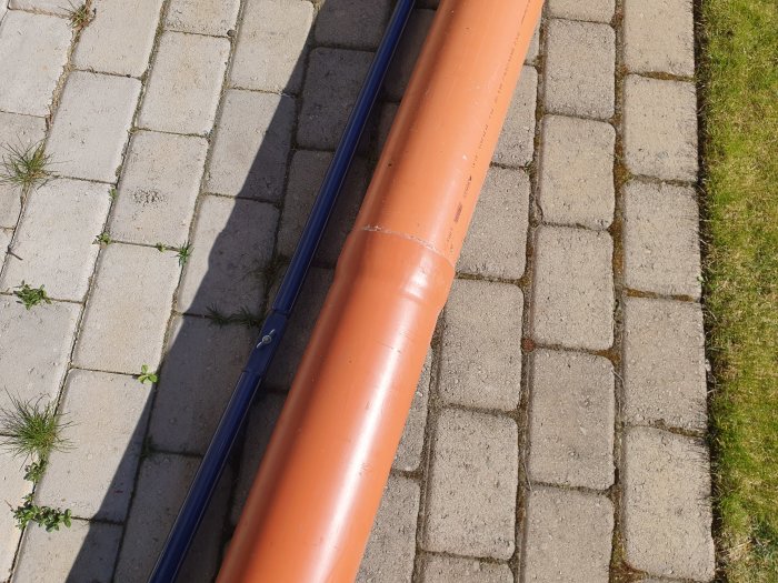 Brunnsrör i orange plast bredvid ett blått rör på ett stenlagt underlag utomhus i solljus.
