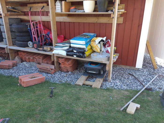 Förvaringsutrymme bakom ett garage med travade däck, säckar med material och verktyg på grusunderlag.