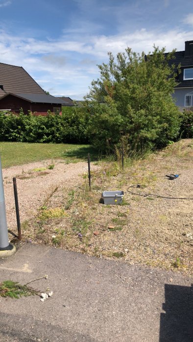 Område under renovering med grus, ogräs och ett borttaget staket, verktyg synligt på marken.