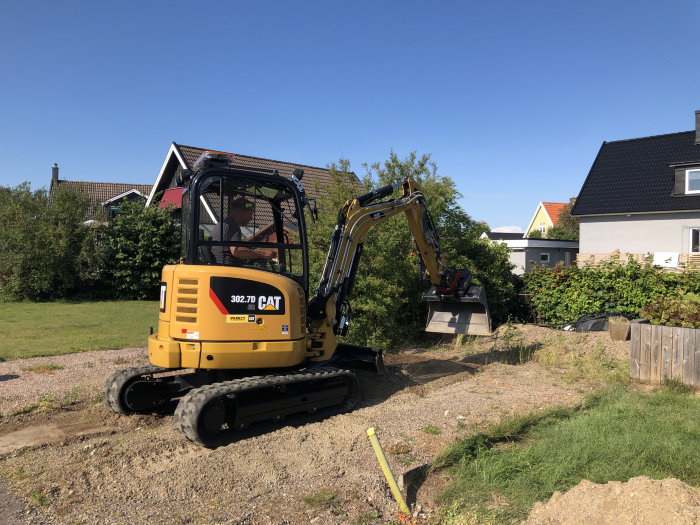 En grävmaskin förbereder marken för stenläggning på en bilplats, med gräs och hus i bakgrunden.