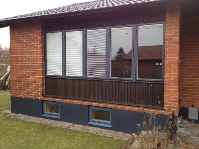 Husfasad med röda tegelväggar, vita fönster med aluminiumutsida och nedre panel i dåligt skick.