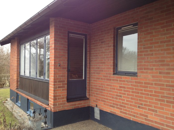 Husfasad med röda tegelväggar, vita fönster omgivna av mörk panel och svarta detaljer.