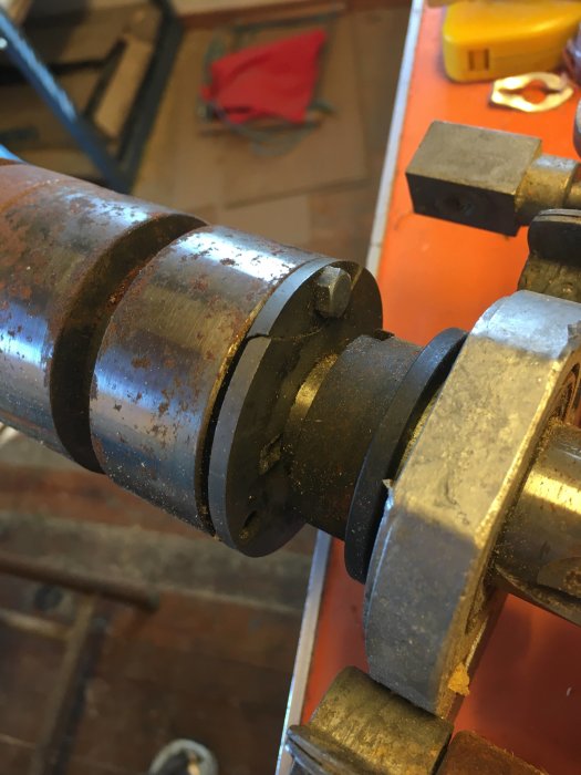 Rostig maskindel med synlig spricka på en svart bricka mellan två cylindriska komponenter i en verkstadsmiljö.