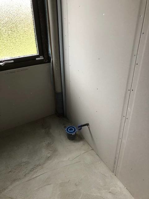 Hörn av badrum under renovering med omonterad gipsvägg och synlig golvbrunn nära fönster.