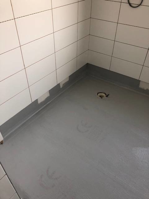 Orenoverat badrumsgolv med synlig golvbrunn och delvis kaklade väggar med plastskydd i hörnen.