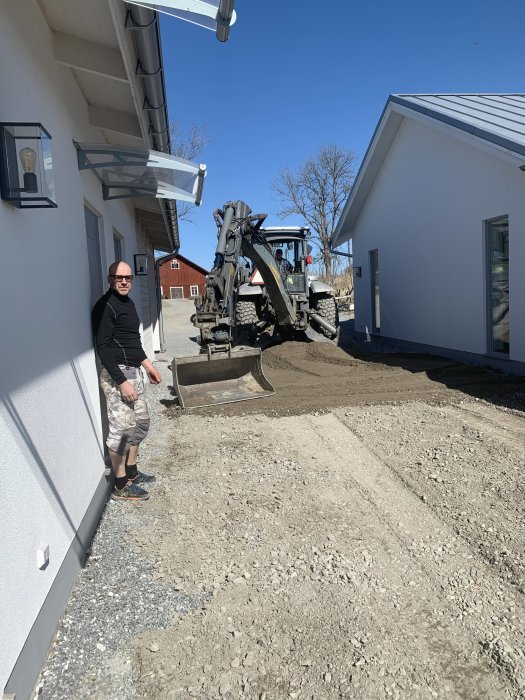 Man lutad mot husvägg bredvid grusad yta med grävmaskin i bakgrunden under en klarblå himmel.