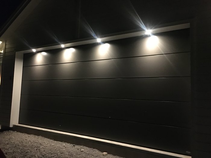 Garageport i silkgrain-yta och NCS-anpassad kulör, belyst av spotlights mot en mörk byggnad.