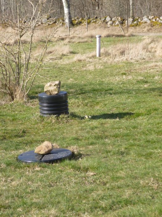 Utsikt över en gräsmatta med ett avloppslock i förgrunden, en fördelningsbrunn i mitten och ett luftrör i bakgrunden.