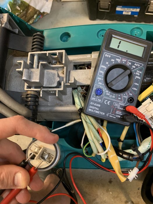 Hand håller mätprober mot en elektrisk komponent, digital multimeter visar avläsning i bakgrunden.