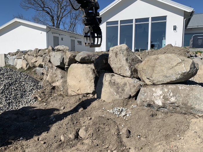 Stenmur under konstruktion bestående av stora stenar med en grävskopa i bakgrunden och en del av ett vitt hus.