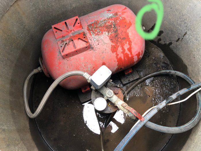 Röd, sliten cistern försänkt i brunnen med kopplade sladdar och slangar, omgivet av vatten och betong.