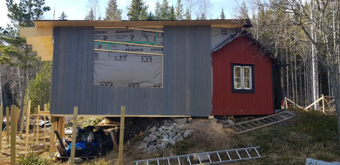 Halvfärdigt utbyggt rött hus med presenningar och byggmaterial omkring i skogsmiljö.