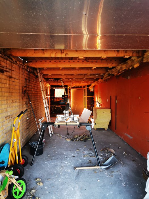 Garage med öppet tak där isolering och gips är borttaget, exponerade bjälklag och byggmaterial med verktyg på golvet.