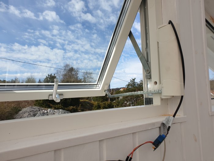 Elektronisk enhet monterad på vägg med lösa kablar framför öppet fönster under en blå himmel.