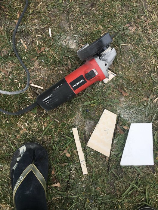 Nedslängd vinkelslip med sladd bredvid krossade kakelplattor och träbitar på gräsmatta, personens skodda fot syns.