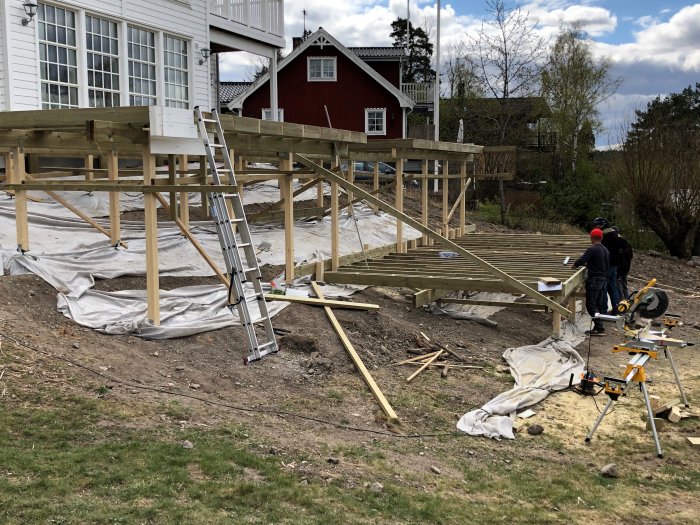 Byggplats för en altankonstruktion med arbetare, halvfärdiga trädäck och träbalkar framför ett hus.