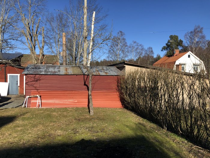 Rött garage med korrugerad plåt och masonitklädda dörrar i en trädgård.