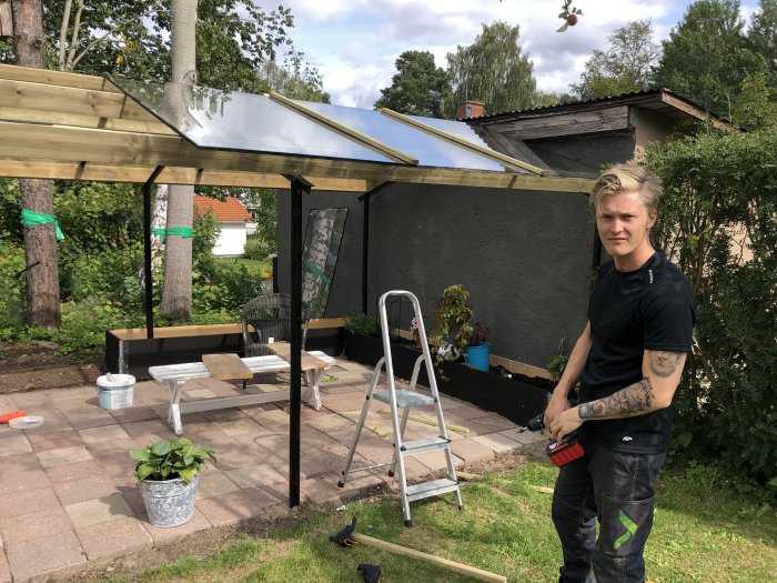 Person bygger pergola med kanalplasttak, markstensgolv och byggmaterial runtomkring.