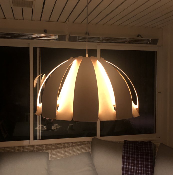 Hängande lampskärm med rundade lameller som lyser varmt i ett mörkt rum med soffa och fönster.