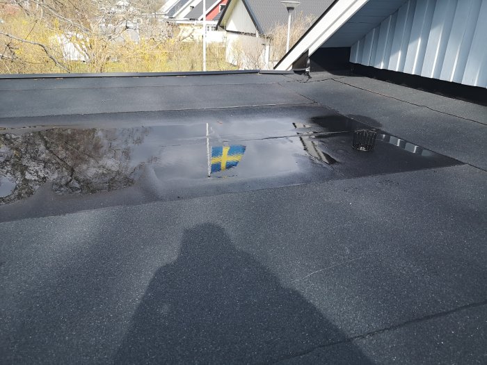 Garagetak med ny lagd papp och vattenansamling runt en högre brunnen, skuggor av personer syns.