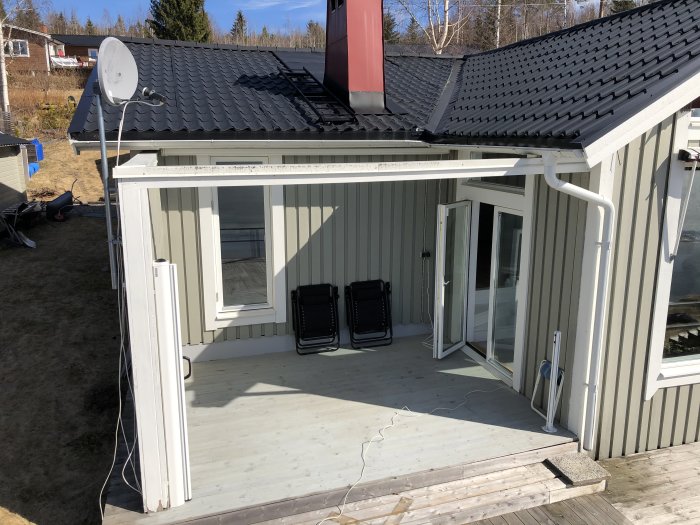 Uterum med öppet skjutparti och sidoglaspartier, på en träterrass bredvid ett hus med satellitantenn.