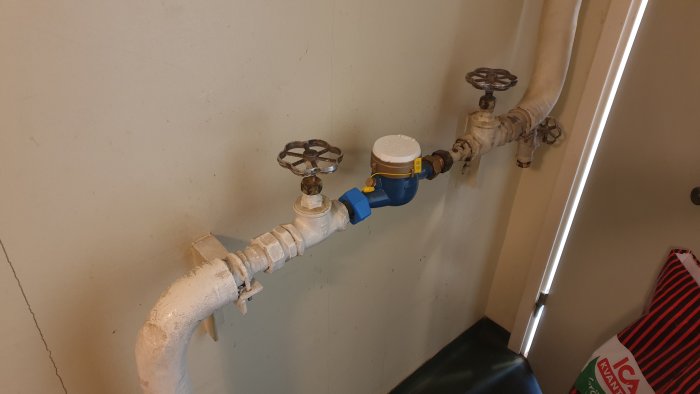 Vattenmätare med huvudavstängningsventil och ett blått reglage mot en vit vägg och rörinstallationer.