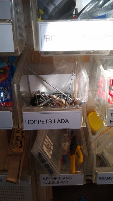 Transparent låda märkt "Hoppets Låda" fylld med olika skruvar bland verktygshyllor.