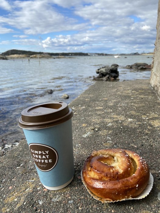 Kaffemugg och kanelbulle på kajkant framför havsutsikt, paus efter trädgårdsarbete.