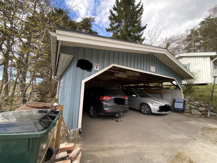 Dubbelcarport med två bilar parkerade under, intill ett bostadshus, med öppna sidor och blå plåtväggar.