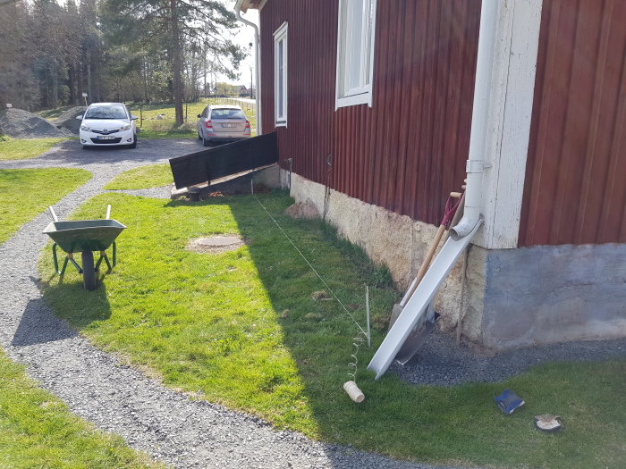 Grävningsarbete vid husvägg med en skottkärra, grävverktyg och påbörjad grävning för fiberslang och vattenledning.