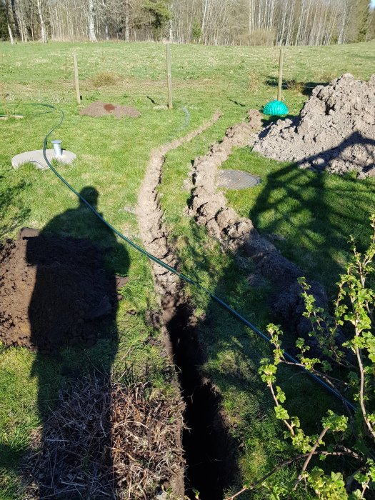 Nygrävd dikesränna för fiberslang och vattenledning i trädgården, med rishög nära grävplatsen.