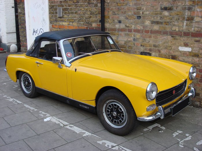 Gul MG Midget parkerad vid tegelvägg, klassisk brittisk sportbil med svart cabriolettak.