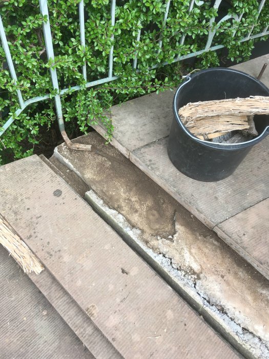 Konkret trappa med löst bruk vid sättstegen, bredvid en hink med brädor, intill en grön häck.