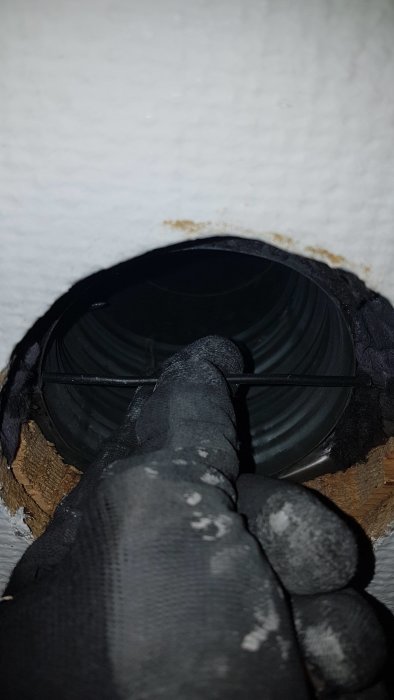 Någons hand med skyddshandskar som håller ett 95-96mm diameter ventilationssrör i ett hål i taket.