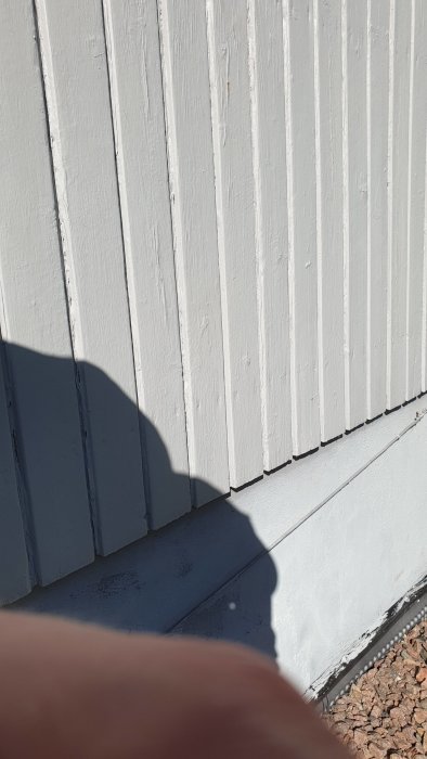 Vertikal ljusgrå fasadpanel med skuggor och en del av en person fotograferad nära panelen.