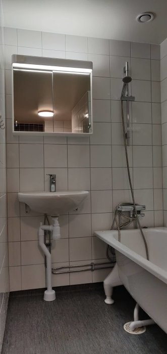 Badrum med vit kakelvägg, handfat, badrumsskåp med belysning och dusch vid sidan av badkaret.