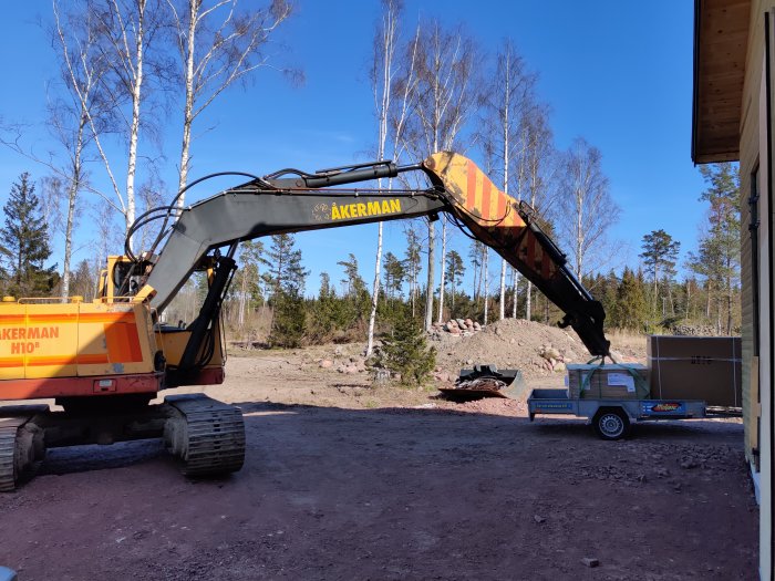 Grävmaskin bredvid en släpvagn med byggmaterial på en tomt i skogsmiljö.