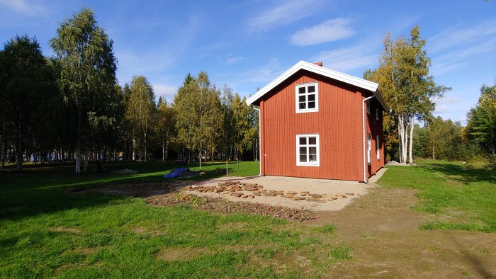 Röd stuga med vita knutar och uteplats under anläggning, järnsand synlig, grön gräsmatta.