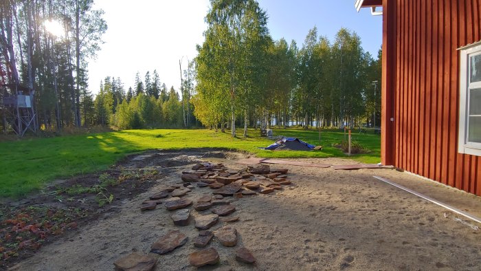 Uteplats under konstruktion med markerad dräneringsledning, sand och stensättning i en trädgård nära en röd husgavel.