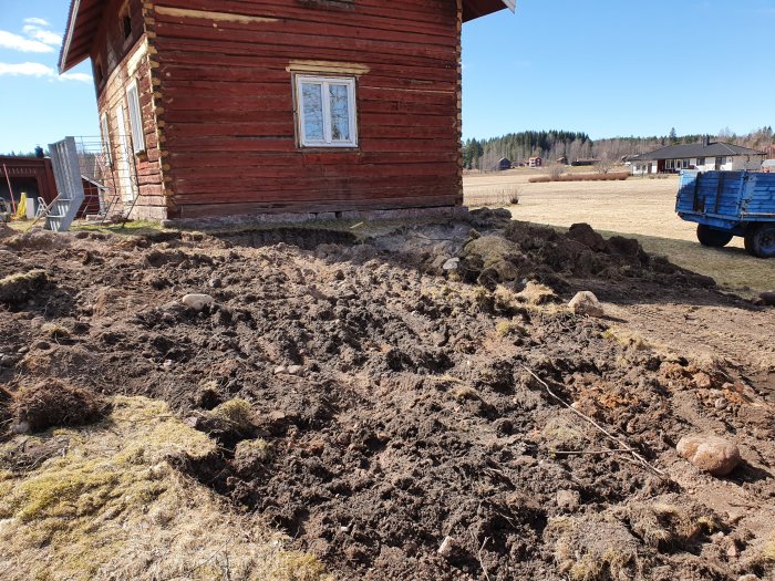 Nygrävd mark vid röd stuga, högar av jord och gräs, landsbygdsutsikt i bakgrunden.