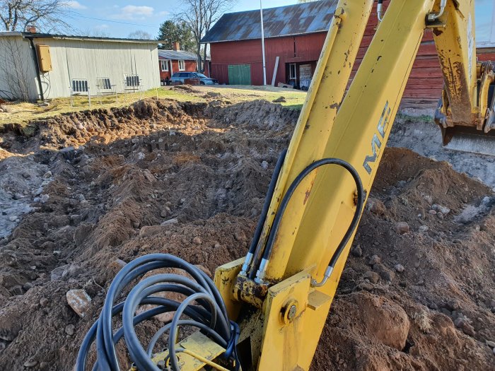 Grävmaskinsarm och hydraulslangar med nygrävd mark och byggnader i bakgrunden.