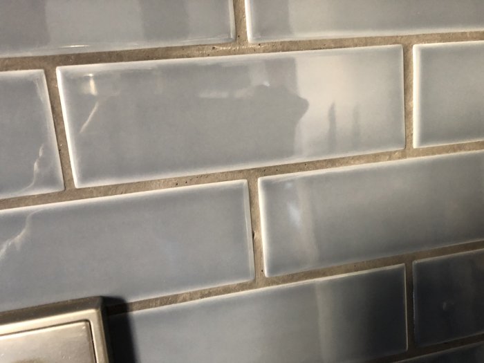 Vägg med kakel vars fogar skiftar i färg från mörkgrå till ljusare nyanser, tecken på felaktig fogning.