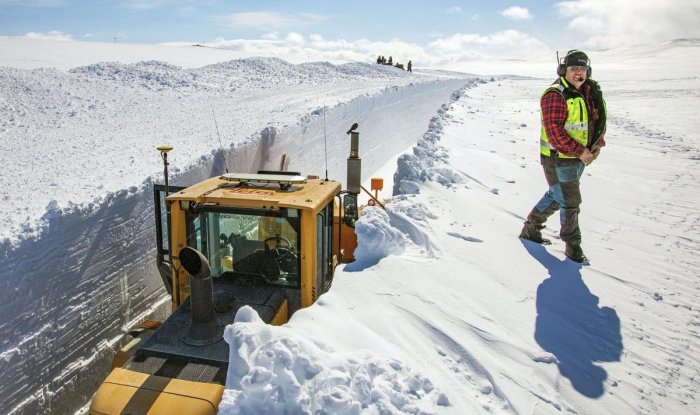 Snöröjningsmaskin och arbetare på snötäckt Vildmarksvägen med 3,5 meter höga snöväggar under blå himmel.