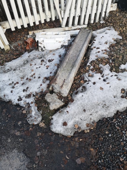 Nedfallen grindstolpe på marken vid vit staket, med snörester och grus.