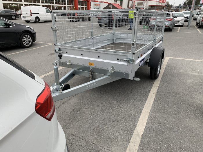Släpvagn med nätgrindar parkerad på parkeringsplats kopplad till vit bil, lastöglor syns på flaket.