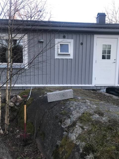 Bild på ett hus med vitt dörr och grå panel, intill en stor bergsknalle som lutar mot huset.