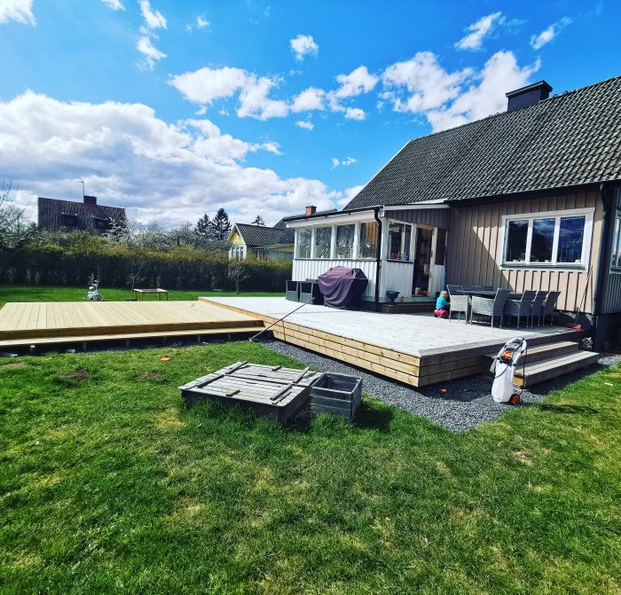 En uteplats vid ett hus med nybyggd trädäck och gräsmatta under en klar himmel med moln.