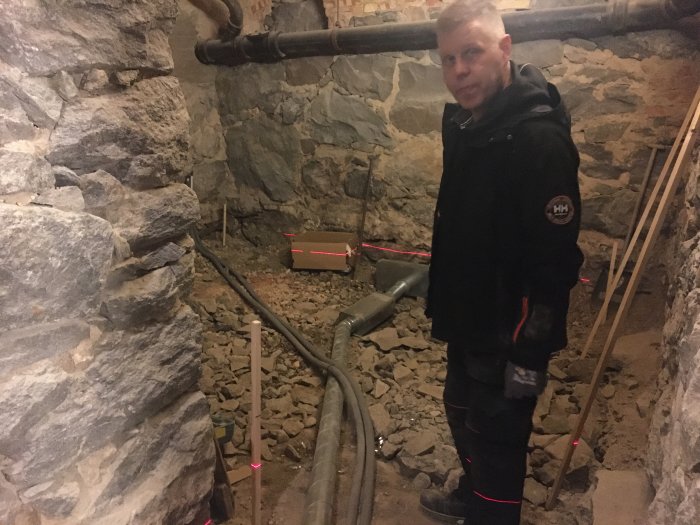 Person står i ett källarutrymme med stenmurar under förberedelser för ventilation och värmegolv.