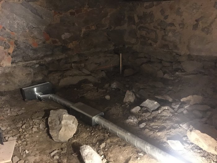 Ventilationsrör ligger på ett grovt grusunderlag i ett outvecklat källarutrymme med stenmurar.
